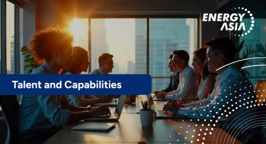 A diverse team of professionals engaged in discussion around a conference table with laptops, backlit by the setting sun in a high-rise office.